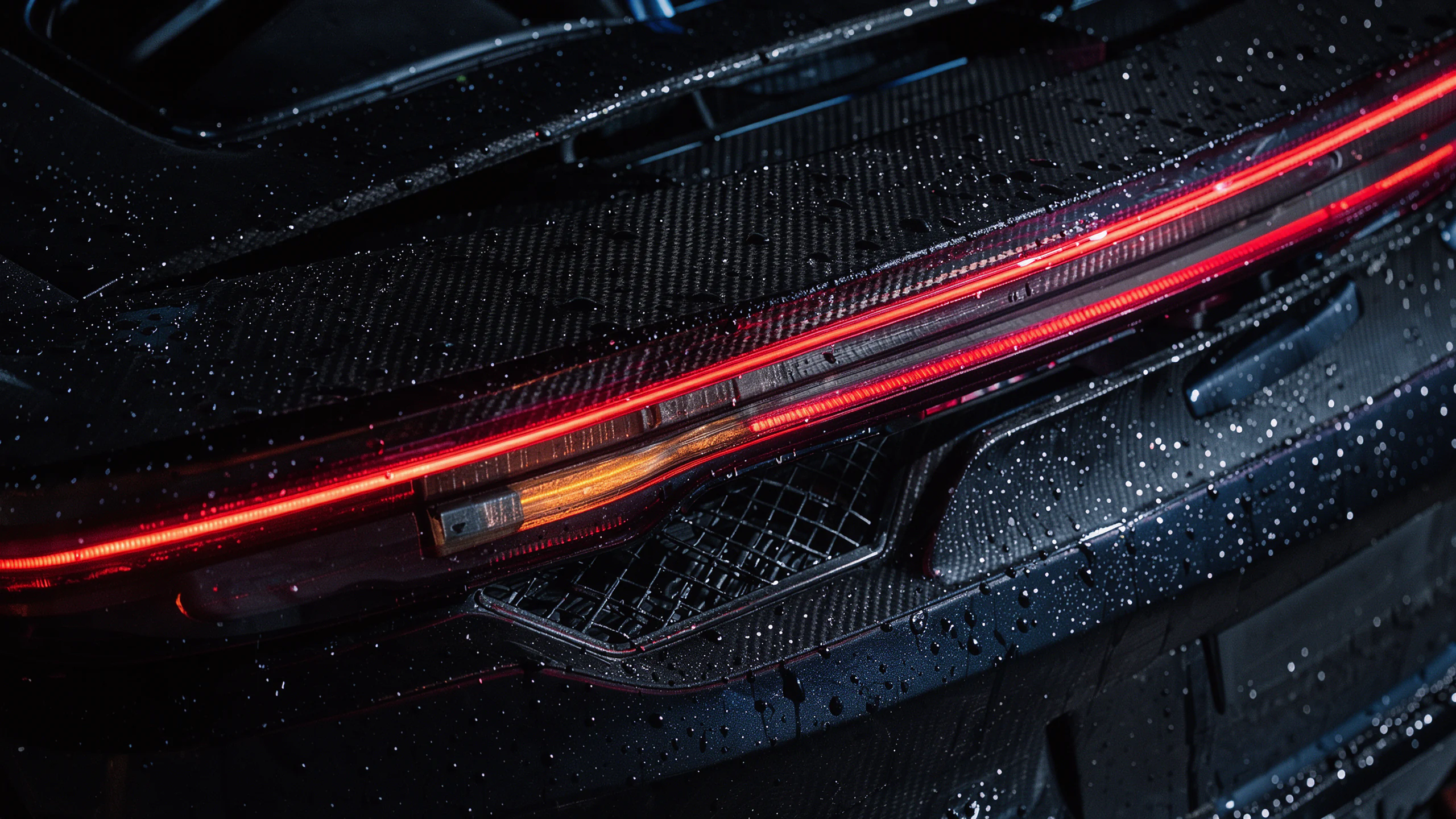 Tail Light — An extreme close-up of the rear light bar and engine vent of a black sports car at night, rain beaded on the bodywork. A thin red LED line glows across the frame, with a faint amber turn signal; the carbon and paint textures are visible under a hard raking light. Composition: macro, diagonal light bar from lower left to upper right, edge-to-edge. Lighting: red practical glow, cool blue rim light, deep blacks. Texture: water droplets, clear coat, carbon weave, metal mesh, fine dust. Mood: precise, expensive, dangerous. Ultra detailed macro automotive photography, photorealistic, no badges, no logos, no text.